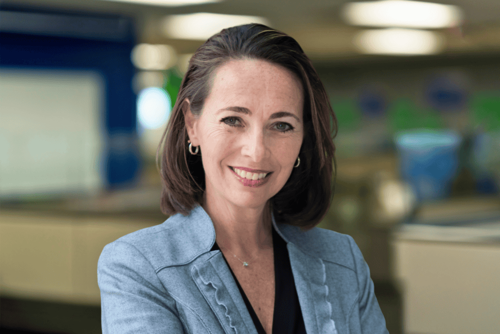 Woman with shoulder-length brown hair in a gray blazer and black top, smiling, standing in an office setting with blurred background.