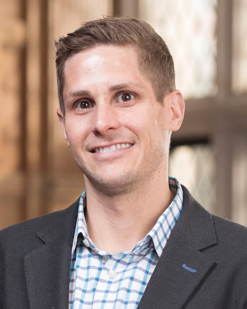 A man with short light brown hair, wearing a checked shirt and dark blazer, stands indoors in front of a blurred background, ready to Get In Touch.