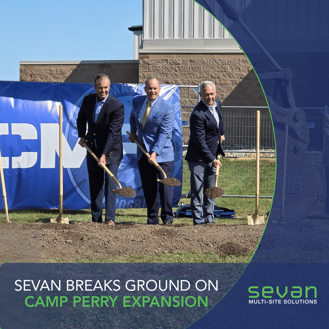 Sevan In The News 1 Three men in suits hold shovels at a groundbreaking ceremony outdoors in front of a blue banner and building, marking the Camp Perry expansion for Sevan Multi-Site Solutions.