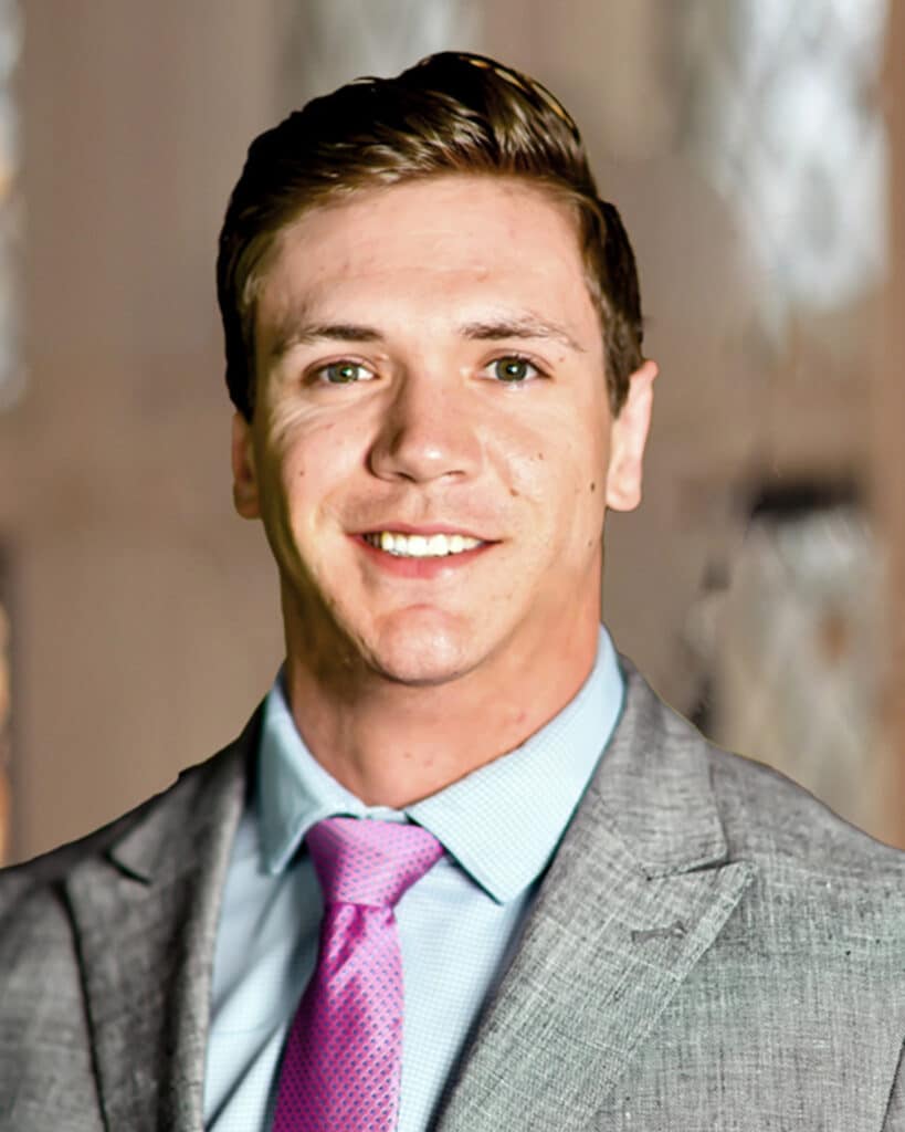 A man in a gray suit jacket, blue shirt, and pink tie is smiling at the camera. The background is softly blurred with neutral colors, creating a welcoming atmosphere that encourages you to get in touch.
