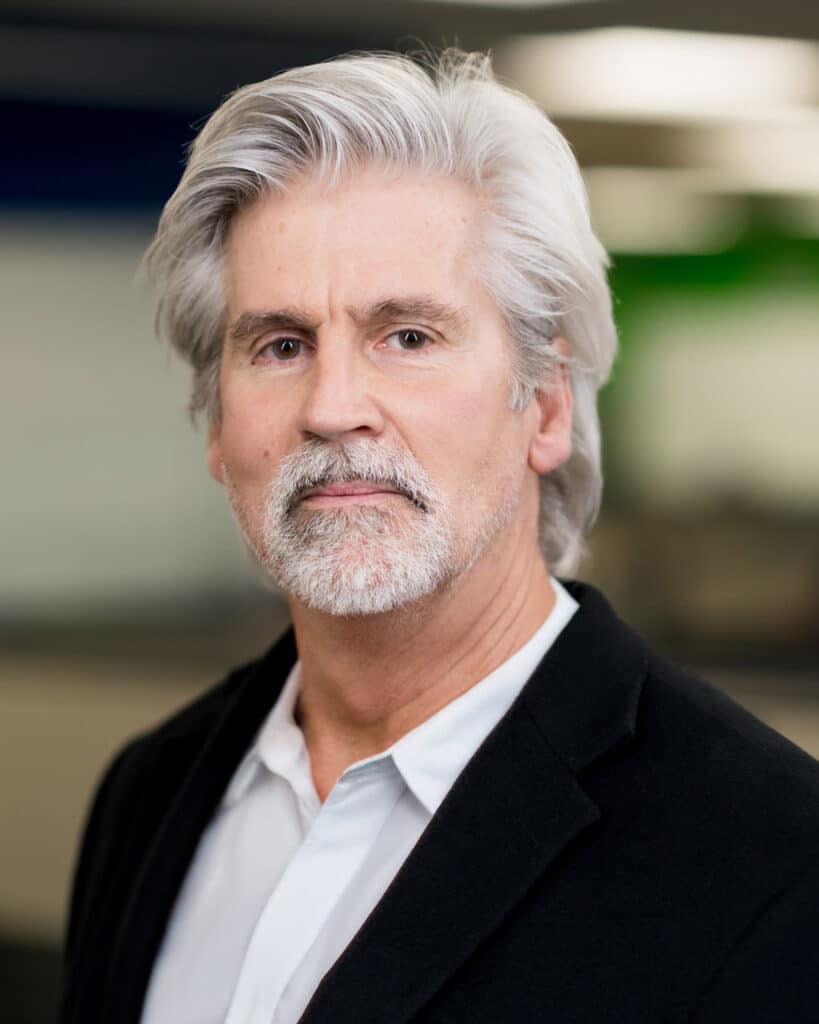 A middle-aged man with gray hair and a beard, wearing a white shirt and dark blazer, stands indoors with a blurred office background, ready to get in touch and assist you.