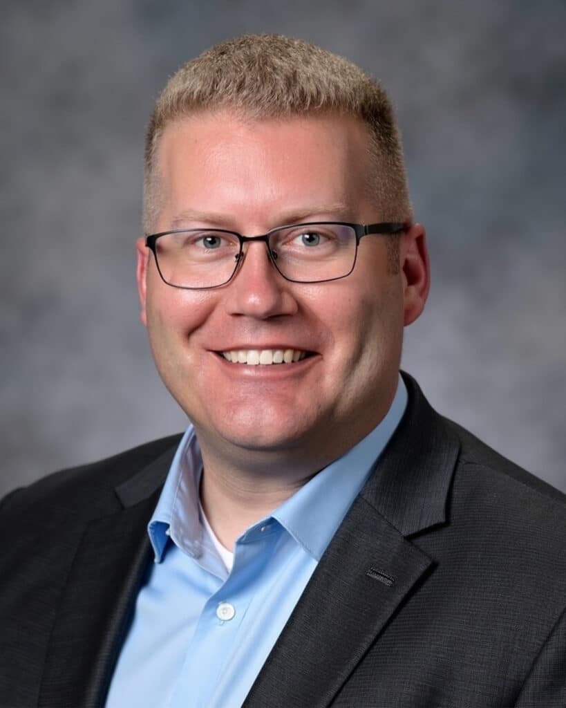 Smiling person with short blond hair, glasses, a light blue shirt, and a dark blazer, set against a gray, blurred background—perfect for anyone looking to get in touch or make contact in a professional setting.
