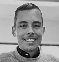 A man with short hair and a trimmed beard, wearing a polka dot shirt under a sweater, smiles at the camera in a well-lit indoor setting.