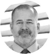 Black and white headshot of a middle-aged man with short hair, a beard, and mustache, wearing a shirt and tie, in front of window blinds.