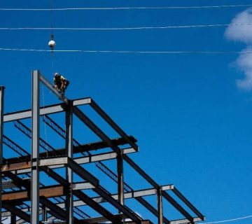 A construction worker sits on steel beams atop a building framework under a clear blue sky, with a crane cable and a few power lines visible overhead.