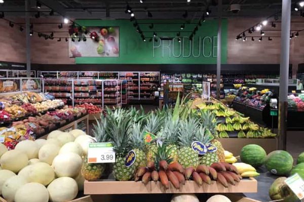 Produce section of a grocery store with displays of melons, pineapples, and other assorted fruits under bright lighting. Signs show prices and product labels.