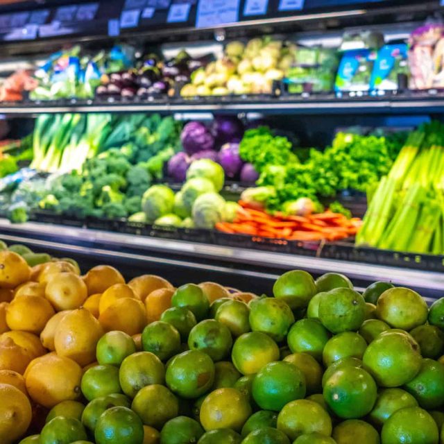A grocery store produce section displays lemons and limes in the foreground, with various vegetables like lettuce, carrots, and eggplant on shelves in the background.