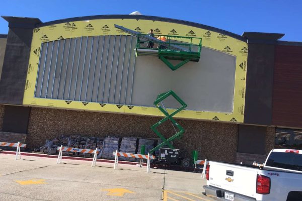 A worker on a green scissor lift installs metal framing on the upper facade of a commercial building under renovation. Construction materials and barriers are visible below.
