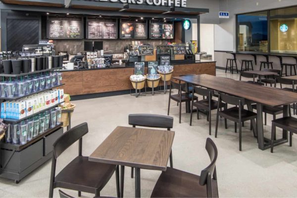 A Starbucks coffee shop interior with wooden tables and chairs, a counter displaying menu boards, and shelves with merchandise and packaged goods.