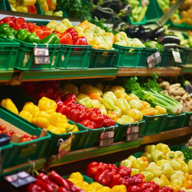 Shelves in a grocery store display a variety of fresh vegetables, including bell peppers, eggplants, and ginger, organized in green plastic crates.
