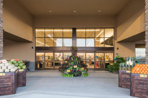 Entrance of a grocery store with large glass doors, fresh produce bins of melons, pineapples, and oranges, and a tiered display of potted flowers and plants.