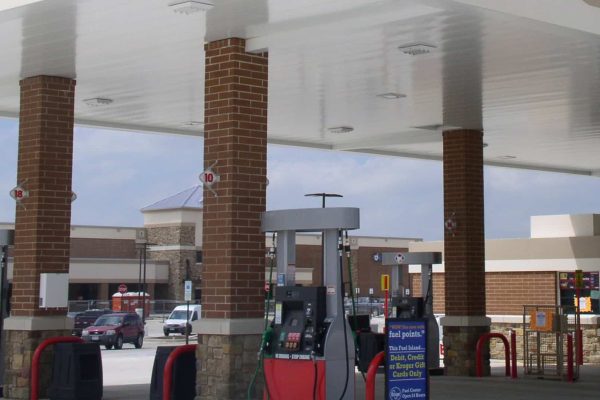 Gas station with multiple fuel pumps under a large canopy, brick pillars, and a convenience store in the background. Several vehicles are parked nearby.