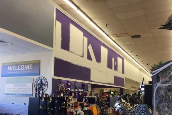 Interior of a retail store with a high wall partially painted purple and white patches, visible ceiling tiles, and various merchandise displayed below.