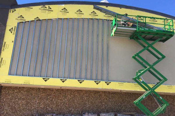 A construction worker on a green scissor lift installs metal wall studs on the exterior of a building under renovation, with yellow sheathing visible.