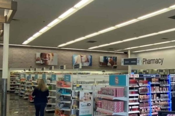 A person walks down an aisle in a brightly lit pharmacy section of a retail store, surrounded by shelves with various products.