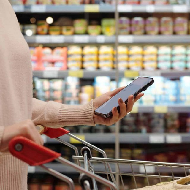 Person holding a smartphone and pushing a shopping cart in a grocery store aisle with shelves of canned goods and food items in the background.