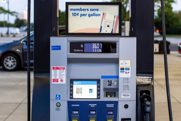 A gas pump at a station displays fuel prices, a digital screen ad for member savings, payment options, and selection buttons. Cars are visible in the background.