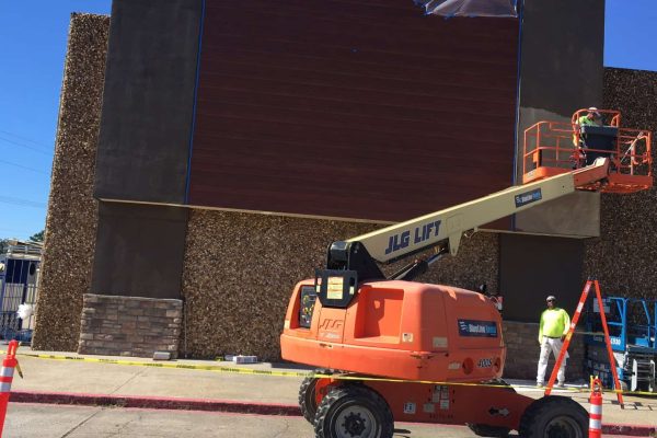 A worker on a JLG lift installs or paints a large sign on the exterior wall of a commercial building, while another person stands nearby. Safety cones and caution tape are visible.