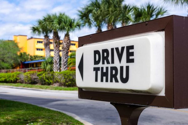 A drive-thru sign with a left arrow stands next to a road lined with palm trees and a bright orange building in the background.