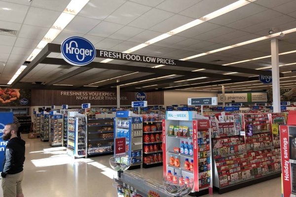 Interior of a Kroger grocery store with aisles of products, greeting cards display, pharmacy section, and overhead signs promoting fresh food and low prices.