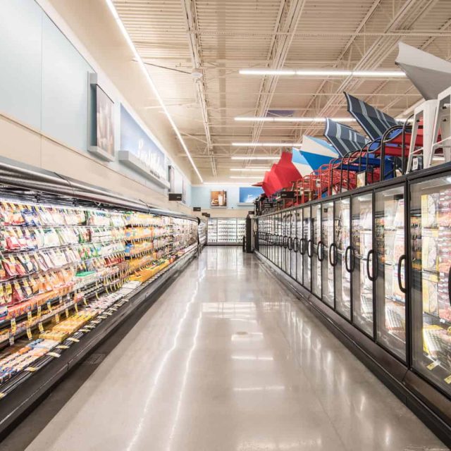 A brightly lit grocery store aisle with refrigerated cases of packaged food on the left and glass freezer doors on the right; metal chairs are stacked on top of the freezers.