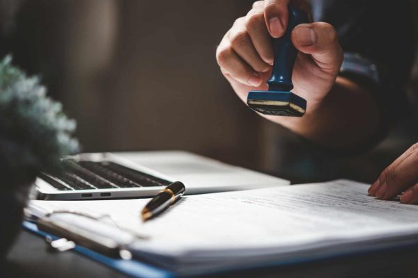 A person stamps a document on a desk next to a laptop and pen.