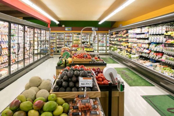 Produce section of a grocery store with fresh fruits and vegetables on display, surrounded by refrigerated aisles stocked with various packaged goods.