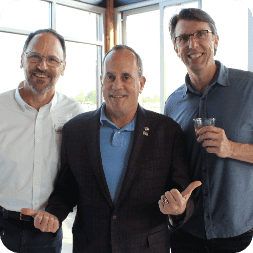 Three men standing together indoors, smiling at the camera; the man in the center is gesturing with both hands while the two men on either side, possibly involved in program management or a co-op internship, wear glasses.