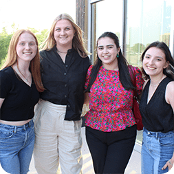 Four young women standing together outside, smiling at the camera, celebrate their internship in construction program management.